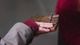 In the centre of this photo pale fingers are stretched out on a wooden board with bits of metal extending out and slightly upwards. The metal colour has a rusted, unpolished look about it. The board is a dark coloured wood, something like a mahogany. The hand looks like it is feeling for information. Just underneath the board in the shadows we see part of another hand, which looks like it is holding the object. The hand feeling the object is partly hidden by a burgundy sleeve cuff which is emerging from a warm dove grey coloured coat sleeve. In the righthand quarter of the image, there is a dark shadow. The top part of this shadow is a similar shade of burgundy as we see in the cuff of the sleeve that is emerging out from the coat.