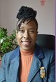 Deborah Husbands: A smiling African Caribbean woman with brown skin. She is sitting in a black chair. Her hair is black with natural locs and styled in an updo with a sideways fringe. She wears a navy-blue jacket with a fabric brooch attached to the right side of the jacket near the lapel. The brooch is multicoloured and in the shape of a small owl. Under the jacket, she wears a terracotta brown polo-neck jumper. To the left of the woman, in the background, is a Jade plant with green leaves. The wall behind the woman is prawn pink with a bright red Chinese symbol in the top right corner.